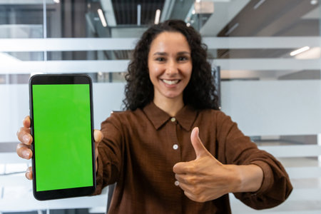 A woman is holding a smartphone with a green screen, smiling and giving a thumbs-up gesture, possibly in an office setting.の写真素材