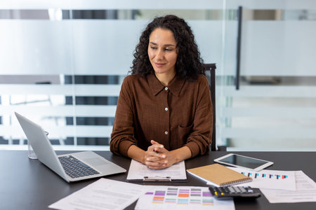 A professional woman is seated at her desk, poised and ready to work in a bright modern office environment. Documents and laptop are present.の写真素材