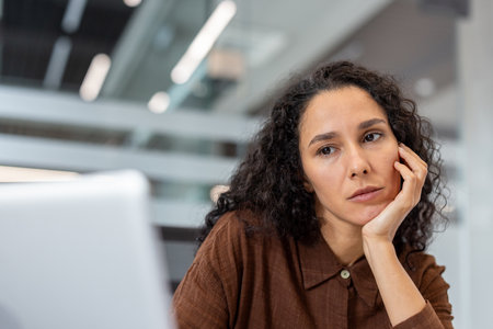 A thoughtful woman rests her head on her hand, gazing pensively into the distance, possibly working on a computer.の写真素材