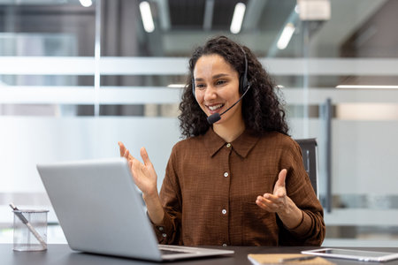 A smiling woman with curly hair wearing a headset communicates via video call from an office, gesturing with her hands.の写真素材
