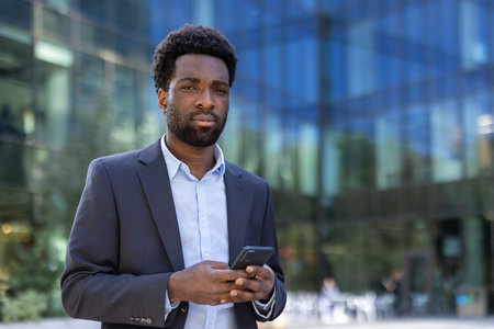 A smartly dressed African-American man holds his smartphone, posing confidently in front of a modern office building.の写真素材