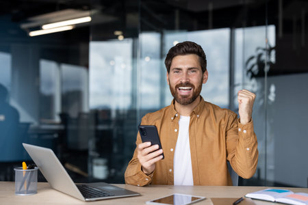 A happy businessman celebrating success while holding a smartphone in an office environment.