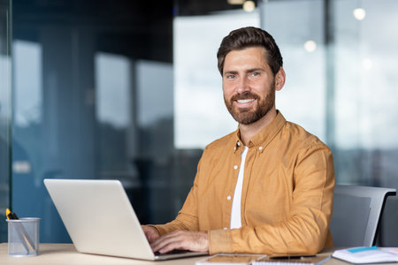 A smiling businessman works on a laptop in a modern office setting, conveying professionalism and...