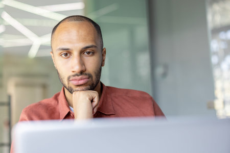 A pensive, dark-skinned man with a beard rests his chin on his hand in front of a laptop, lost in...