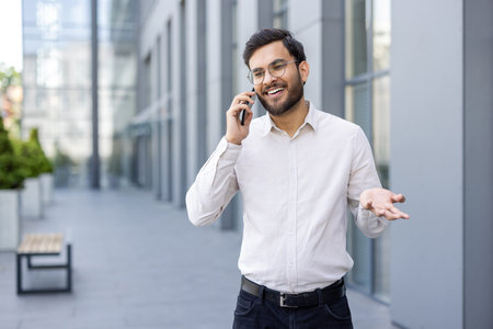 A smiling businessman talks on his phone in front of a modern building. He gestures with his hand while discussing something.の写真素材