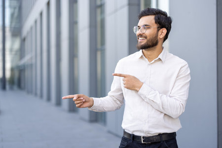 A smiling businessman of Indian descent points sideways, wearing glasses and a button-down shirt, outdoors with a modern building in the background.の写真素材