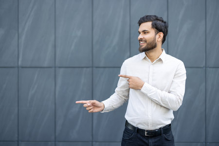 A smiling man in a white shirt points to the side against a backdrop of a gray tiled wall.の写真素材