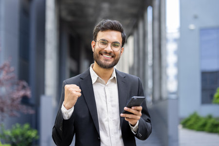 A smiling businessman in a suit holds a phone, celebrating success outdoors with a clenched fist.の写真素材