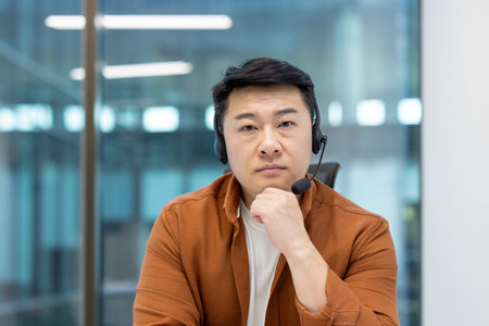 Asian man wearing a headset, looking focused, with his hand on his chin, in a modern office setting.の写真素材