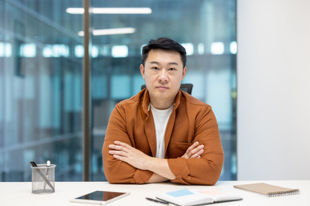 An asian man in a brown shirt and white t-shirt sitting at a desk with arms crossed.の写真素材