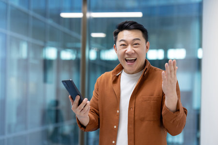 An Asian man, smiling broadly and gesturing with excitement while holding a smartphone, appears pleased with the content.の写真素材