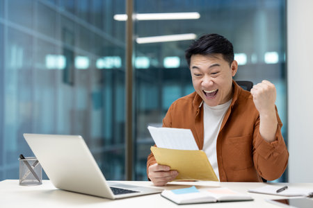 An excited Asian man celebrates with a raised fist after reading good news from a letter. Hes sitting at a desk with a laptop.の写真素材