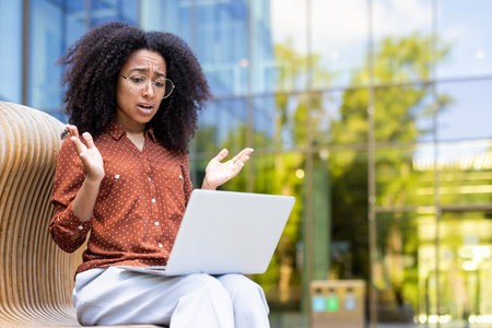 A surprised woman with glasses looks at a laptop. She sits outside, possibly working or video chatting.の写真素材