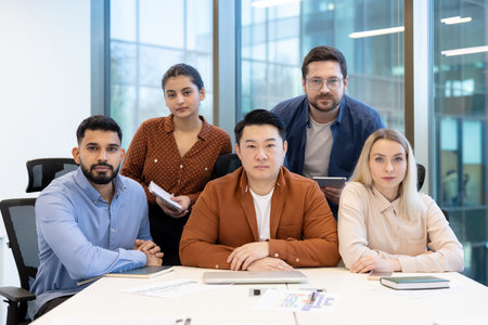 A diverse team of colleagues pose together at a table in a modern office setting, looking directly at the camera.の写真素材