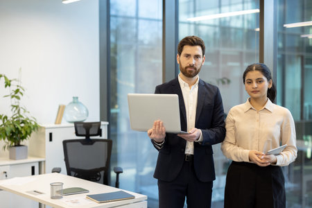 A man and woman in an office setting, the man holding a laptop and the woman a tablet, both looking forward.の写真素材