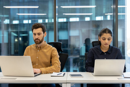 Two diverse colleagues work diligently at their laptops in a modern office, focused on their tasks.の写真素材