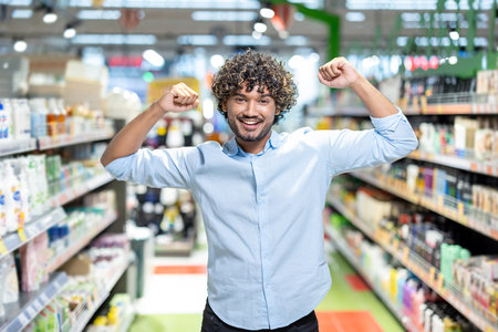 A happy man with curly hair in a supermarket raises his arms in celebration, surrounded by shelves.の写真素材