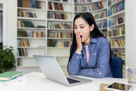 A woman yawns while working on a laptop at a desk in a library, surrounded by bookshelves.の写真素材