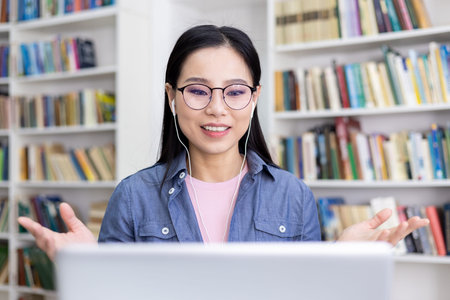 An Asian woman smiles while participating in a video call, using earphones, with a library in the...