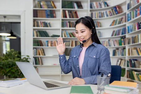 Asian woman in a library using a laptop, wearing a headset, and waving during a video call, with...