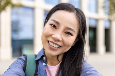 A young Asian woman smiles brightly, taking a selfie with a building in the background, conveying joy and positivity.の写真素材