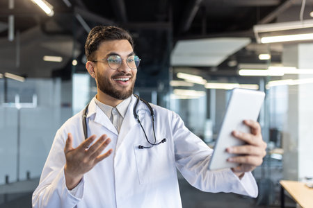 A smiling doctor in a white coat holds a tablet, gesturing during a video call in a modern office...