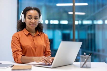 A smiling woman wearing glasses and headphones sits at a desk, working on a laptop in a modern office setting.の写真素材