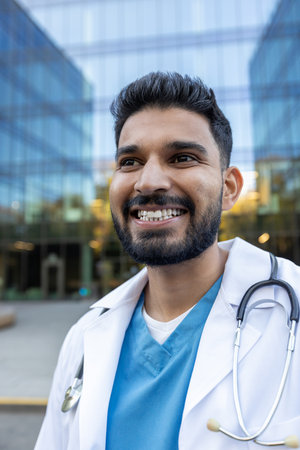 A smiling male doctor with a stethoscope looks away from the camera near a glass building, during daytime.の写真素材
