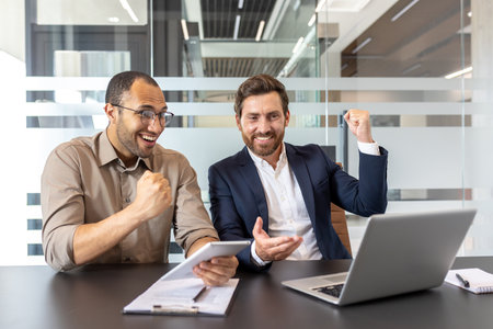 Two business men celebrating a win, one holding a tablet, the other a laptop, both look very happy.の写真素材