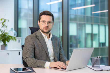 Portrait of a serious and confident young man in a suit and glasses sitting in the office at a desk, working on a laptop and looking at the camera.の写真素材