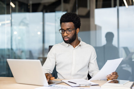 An African American man in glasses sits at a desk in an office, working on a laptop and reviewing documents.の写真素材