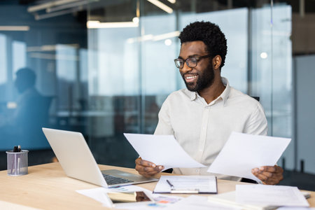 An African American man smiles while reviewing documents at his desk in a modern office setting, next to a laptop.の写真素材