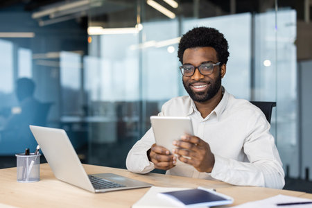 A smiling African American man in glasses uses a tablet at his office desk, a laptop sits nearby.の写真素材