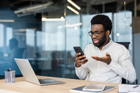 An African-American man wearing glasses is displeased with the information on his phone while working in his office.の写真素材