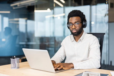 A focused man wearing glasses and headphones works on a laptop in a modern office setting, looking directly at the camera.の写真素材
