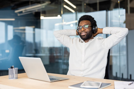 A relaxed African American man wearing headphones and glasses smiles at his desk in a modern office setting.の写真素材