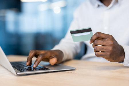 An individual, with dark skin, is using a laptop and holding a credit card, possibly for online payment or a transaction.の写真素材