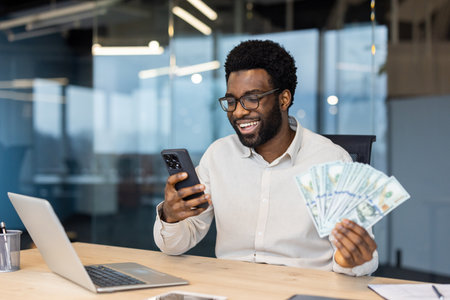 A smiling Black man in glasses holding money and looking at his phone, likely at good news in an office setting.の写真素材