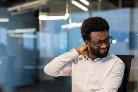 A man with glasses touches his neck, appearing to suffer from neck pain or strain while sitting in the office.の写真素材