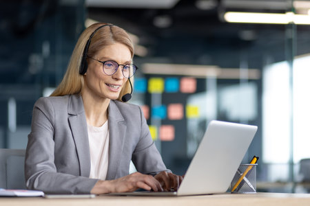 A businesswoman in a headset works on a laptop in a modern office setting, smiling as she types.の写真素材