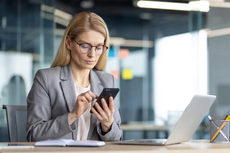 A businesswoman in a modern office setting, focused on her smartphone. Shes wearing glasses and a suit, working at her desk.の写真素材