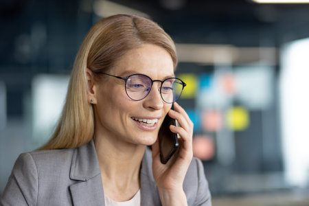 A smiling businesswoman with blonde hair wears glasses while speaking on a cell phone, likely conducting business in an office setting.の写真素材
