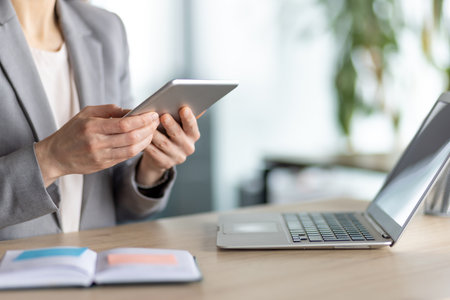 A businesswoman uses a tablet, with a laptop and notepad on a wooden desk. The office environment is blurred in the background.の写真素材