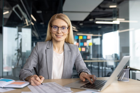 A smiling businesswoman in a modern office setting, working on a laptop while taking notes, radiating confidence.の写真素材