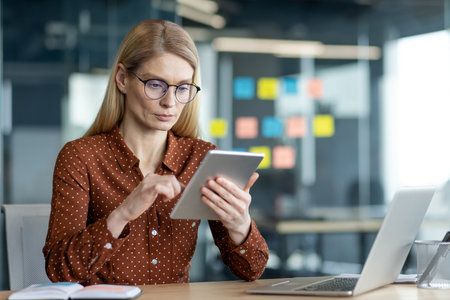A businesswoman is working at her desk with a tablet and laptop in a modern office setting, wearing glasses and a button-down shirt.の写真素材