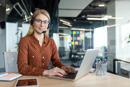 A smiling blonde woman wearing a headset works on a laptop in a modern office setting. She looks friendly and helpful.の写真素材