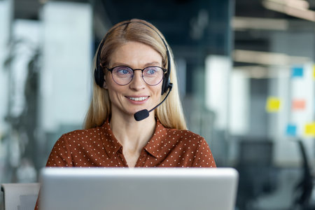 A smiling blond woman wearing a headset and glasses works at a laptop in a modern office setting, possibly customer service.の写真素材