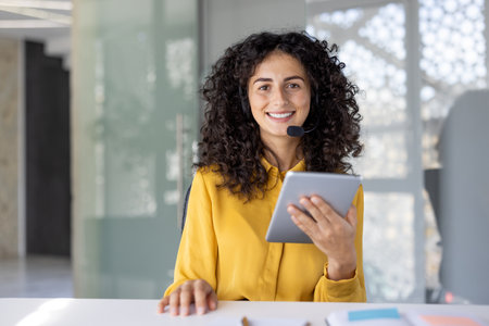 A smiling woman with curly hair wearing a headset, holding a tablet, looks directly at the camera in an office setting.の写真素材
