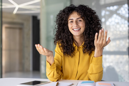 A cheerful woman with curly hair, gestures expressively while smiling, possibly presenting or explaining something in an office setting.の写真素材