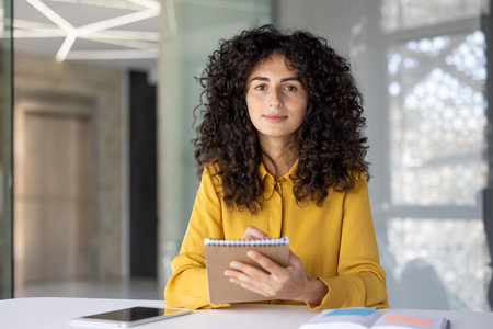 A woman with curly hair sits at a table, holding a notebook, likely for an online presentationの写真素材